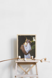 Oak framed canvas print of a woman holding a baby resting on a wooden stool against a white wall