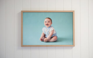 Oak framed canvas print of a baby sitting on a blue backdrop hung on a white vj groove panelling wall