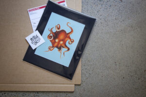 Black matted art print of octopus in plastic sleeve sites on carboard packaging with postage label and thank you card ready for drop shipping