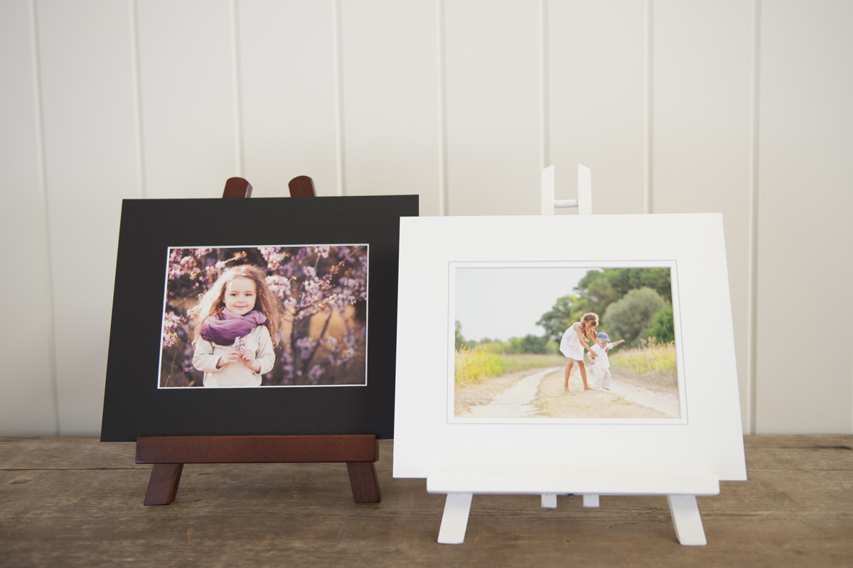 Two matted prints on a wooden bench, one has a black matt with an image of a girl resting on a wooden easel photo stands. The other is a white matted print of a girl and boy sitting on a white easel