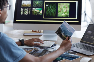 Back view portrait of young African-American man holding printed photographs while using editing software via computer while working at desk in home office