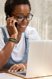 Stylish African American woman calls on smart phone looks in laptop computer, updates software, has phone conversation